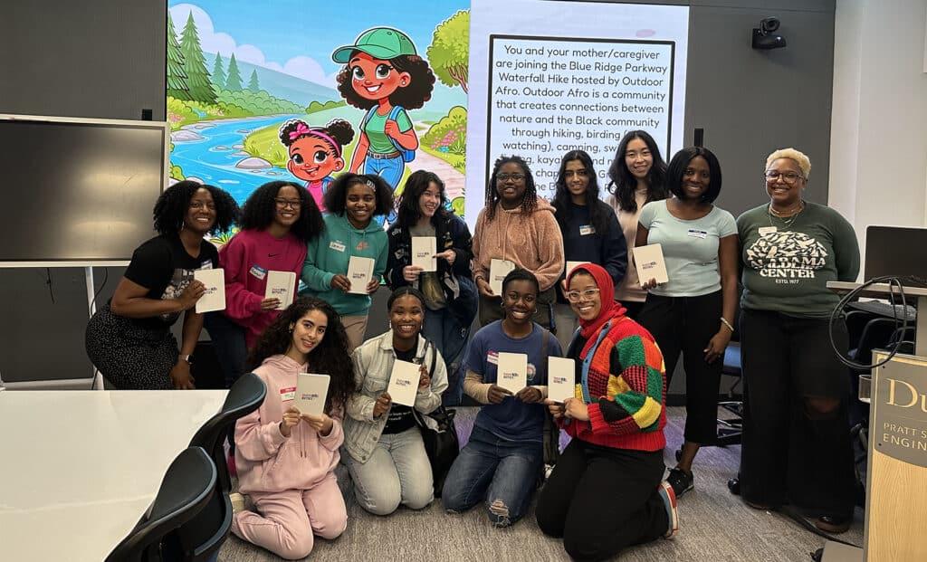 A group of students posing together in a classroom during a research activity, holding small booklets, with a colorful presentation slide displayed behind them.