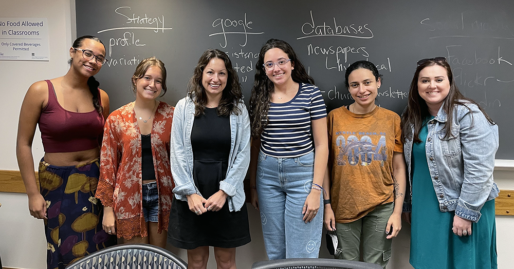 Six young women stand in front of a chalkboard in a classroom.