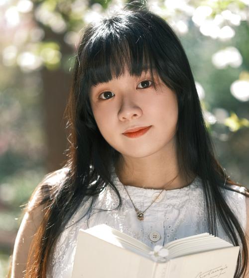 Headshot of Finnie Zhao in a white outfit reading a white book with out of focus trees behind her.